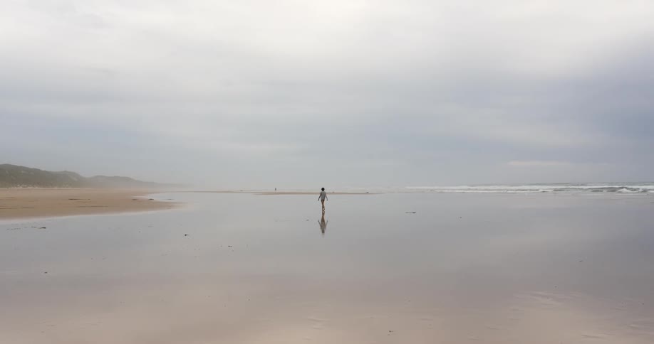 Teenage boy walks around on the wet sand beach next to the ocean on a cloudy day