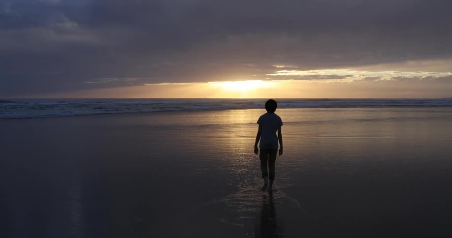 Teenage boy walking barefoot on the wet sand beach is silhouetted against the sunset