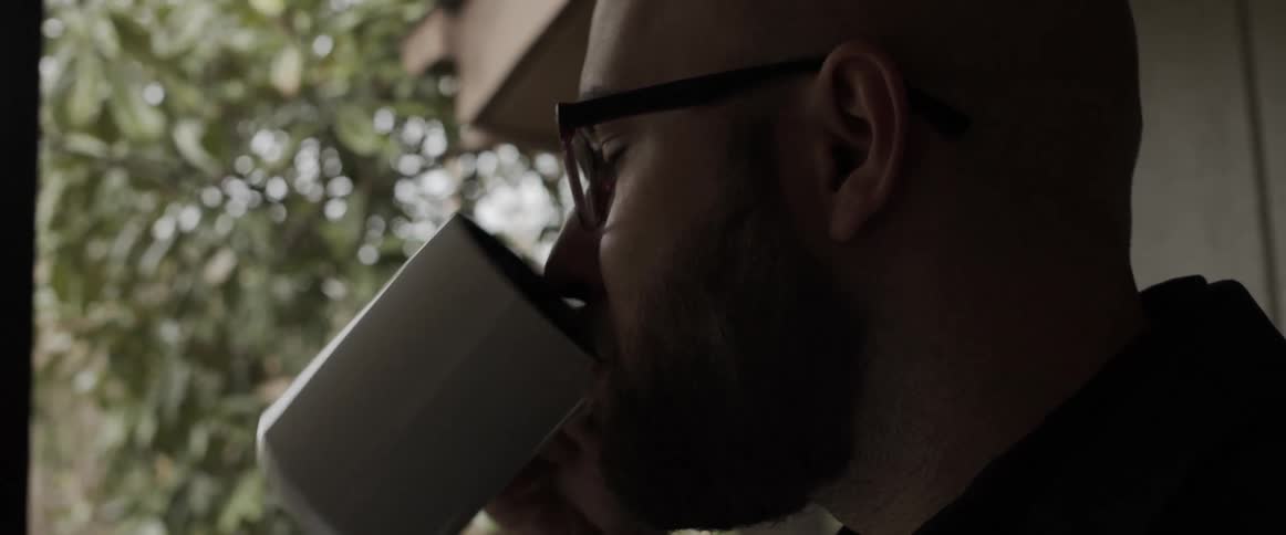 A man in a room with glasses drinking from a mug with a wall in the background