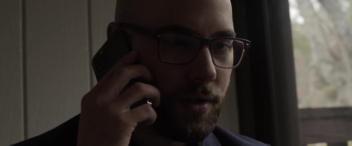 A man speaks on a smartphone while standing near a window indoors during the day