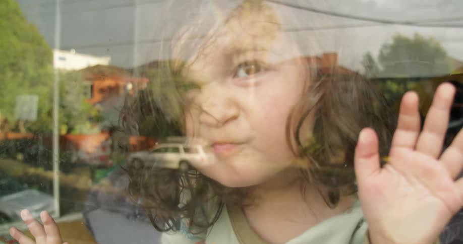 A child pressing her face on a glass window with the reflection of trees houses the sky and clouds on it