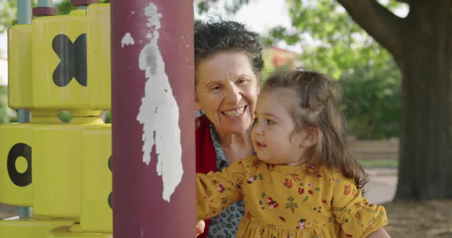 Grandmother smiles as granddaughter plays with tic tac toe game in playground near street