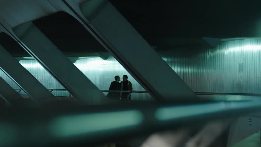 Two men kiss as they walk side by side through an empty terminal