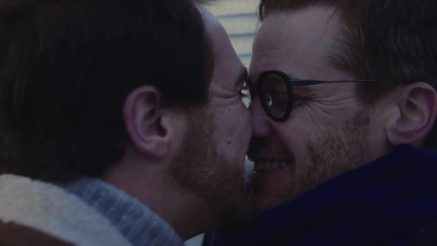 A pair of happy men kiss one another while standing outdoors in a city in winter clothing in the evening