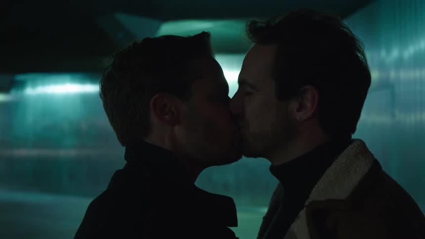 Two men press foreheads together then kiss near wall reflecting overhead light