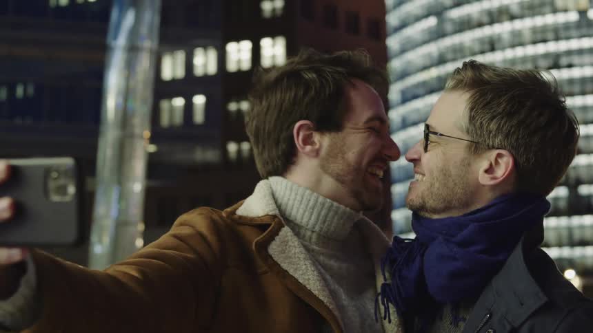 A male couple smiling and kissing while taking a selfie in the city at night