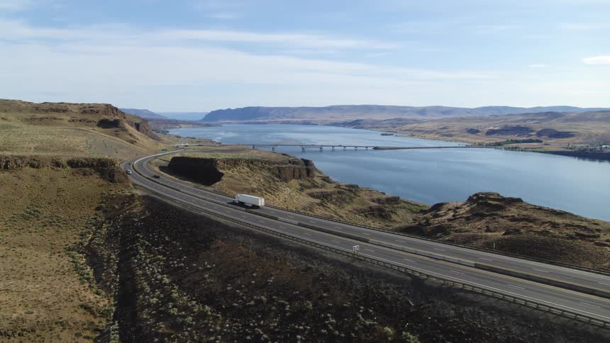 Cars and trucks drive on a steep grade along a highway that leads down to a river