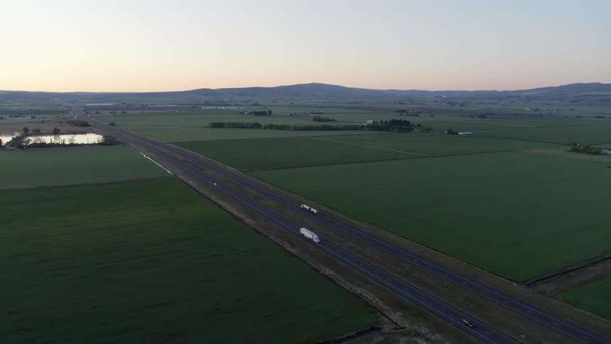 Cars and trucks travel on a divided highway through lush farm land with hills on the horizon