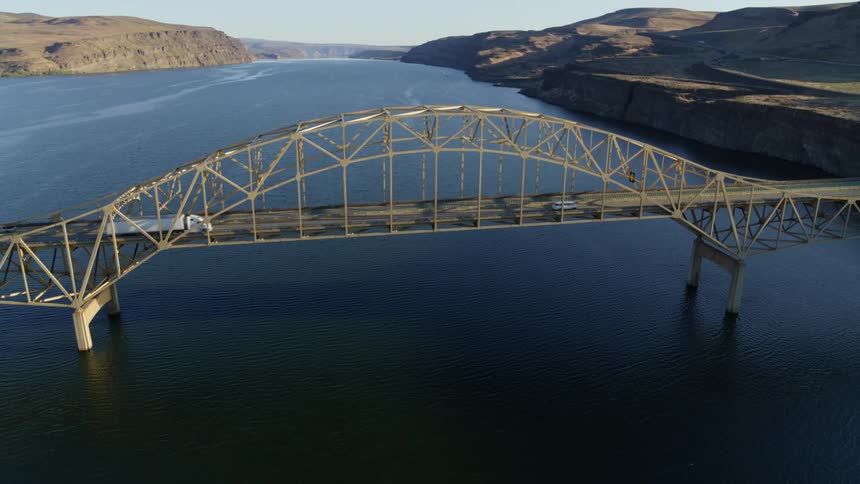 Big rigs travel across a bridge spanning a large river surrounded by mountain foothills