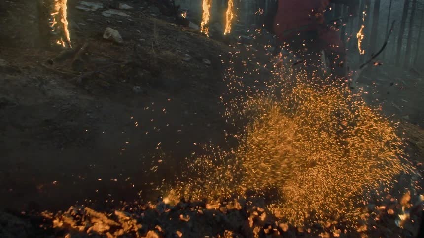 A man on a bike in a forest with burning trees rides through ashes on the ground and scatters embers into the air