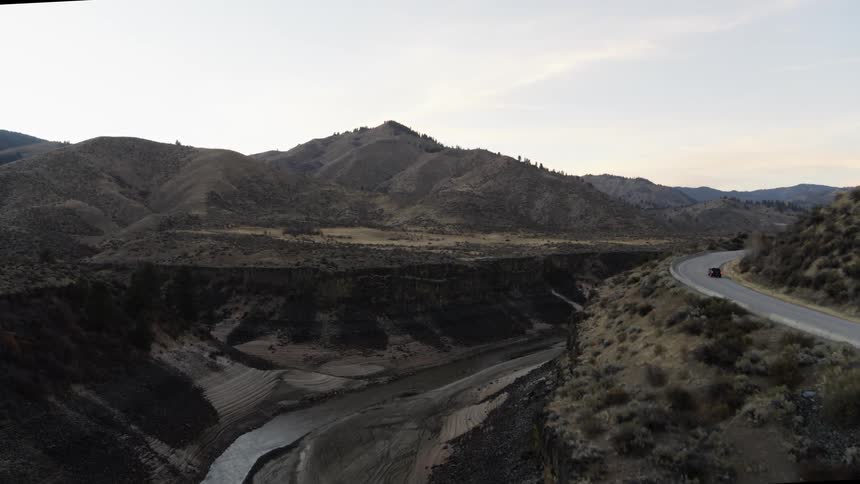 A black pickup truck drives on a curved road near a deep river gorge with foothills in the distance