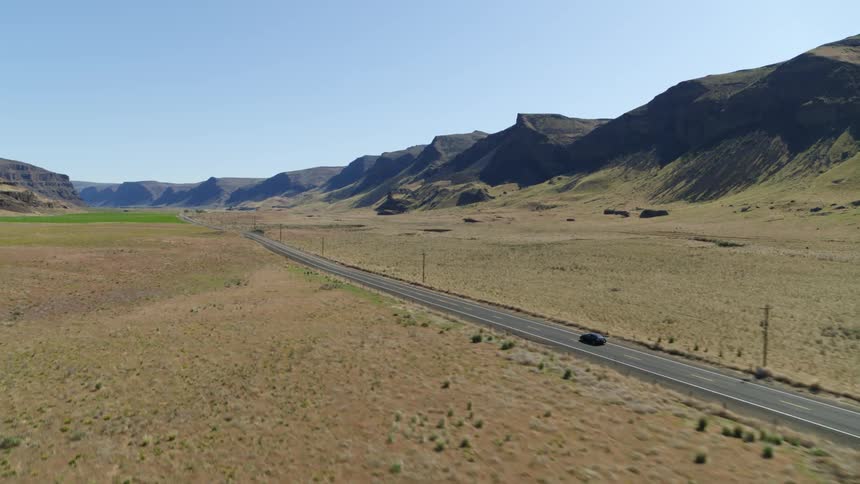 Tracking ascent behind a black car driving along an empty road through a dry canyon valley