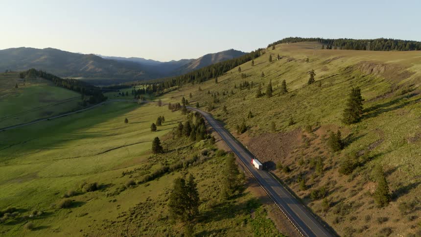 A semi truck drives along a beautiful hillside road towards mountains in the distance