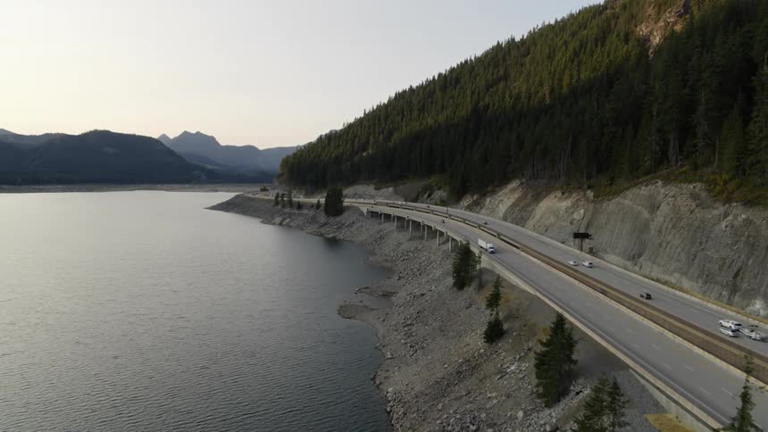 Vehicles driving on a highway along the edge of an alpine lake beween mountains and forests