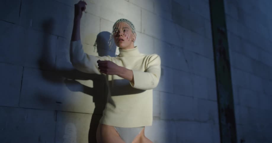 A woman dances while leaning against a cinderblock wall wearing a leotard and beaded headpiece