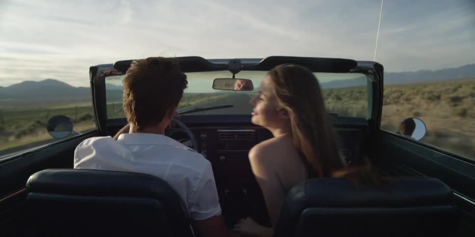 A man and a woman driving a convertible in a desert road with the sky and clouds in the background