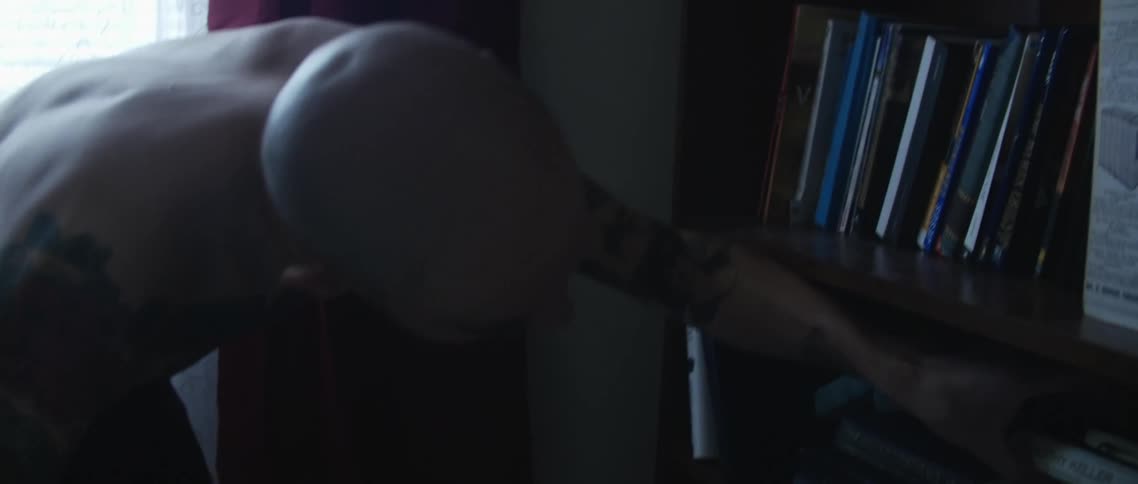 A man with tattoos on his chest and arms searches a bookcase