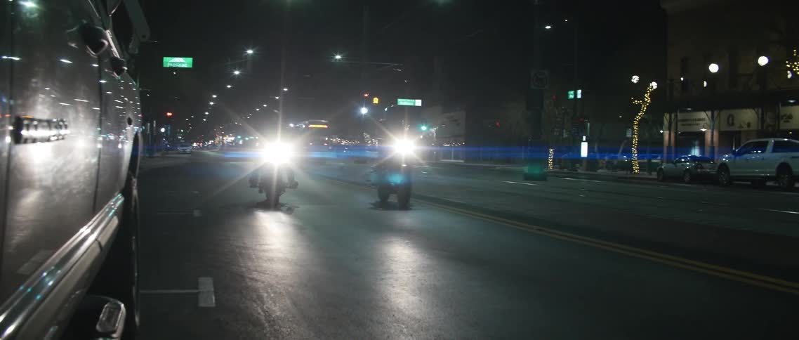 Two people on motorcycles with headlights riding on a city street with buildings in the background