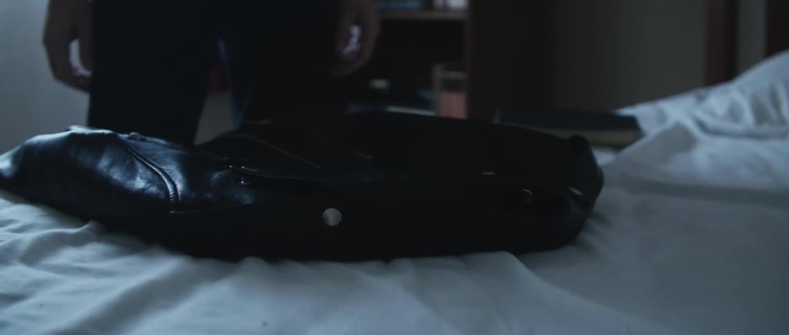 A shirtless tattooed man puts a black leather jacket over the bed in his bedroom