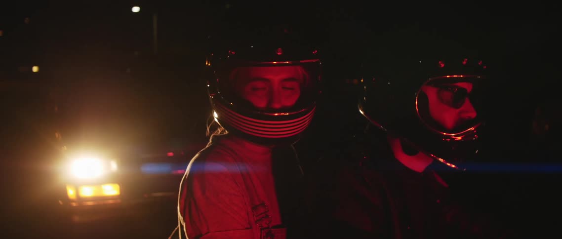 Two people with helmets on a motorcycle with a vehicle with headlights in the background