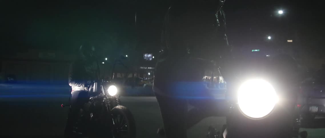 A person with a jacket and a helmet sitting on a motorcycle with headlights with a vehicle in the background