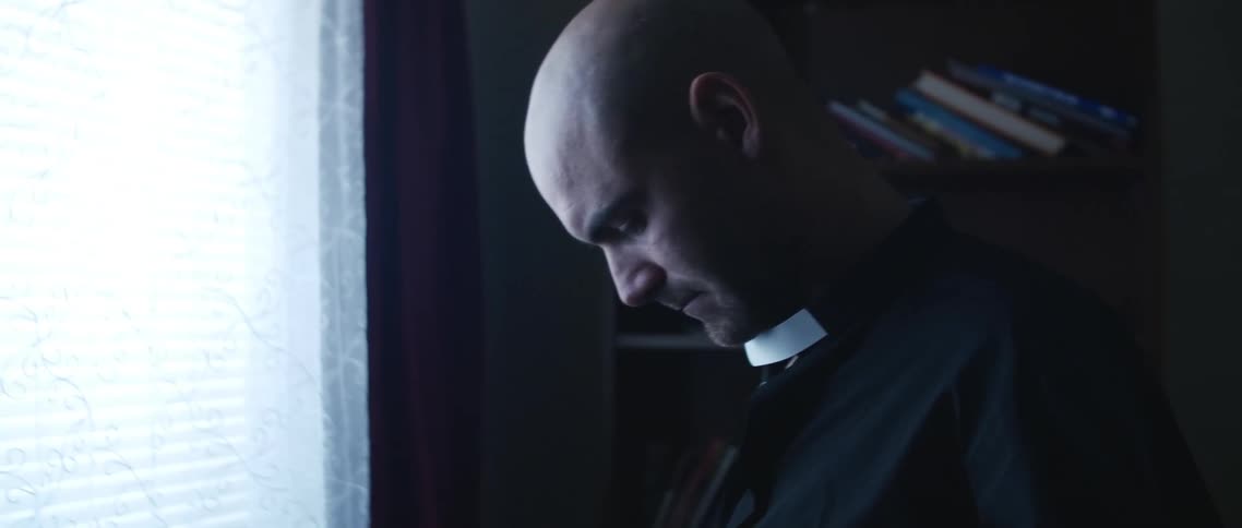 A man in a room next to a window buttoning up his shirt with a bookshelf in the background