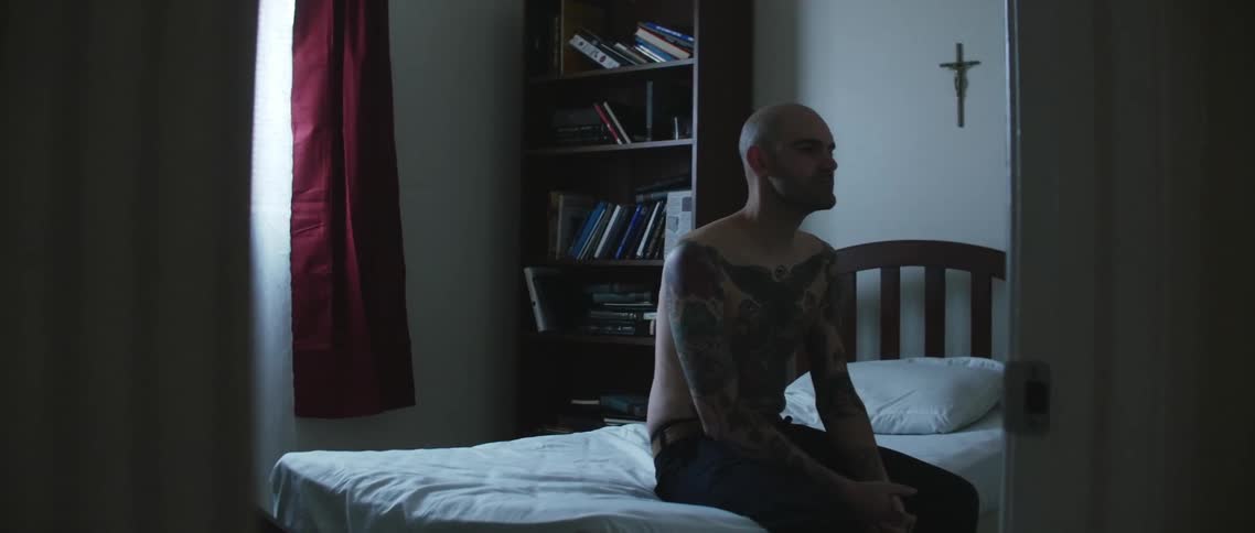 Shirtless tattooed man sitting in bed at home looking at the wall