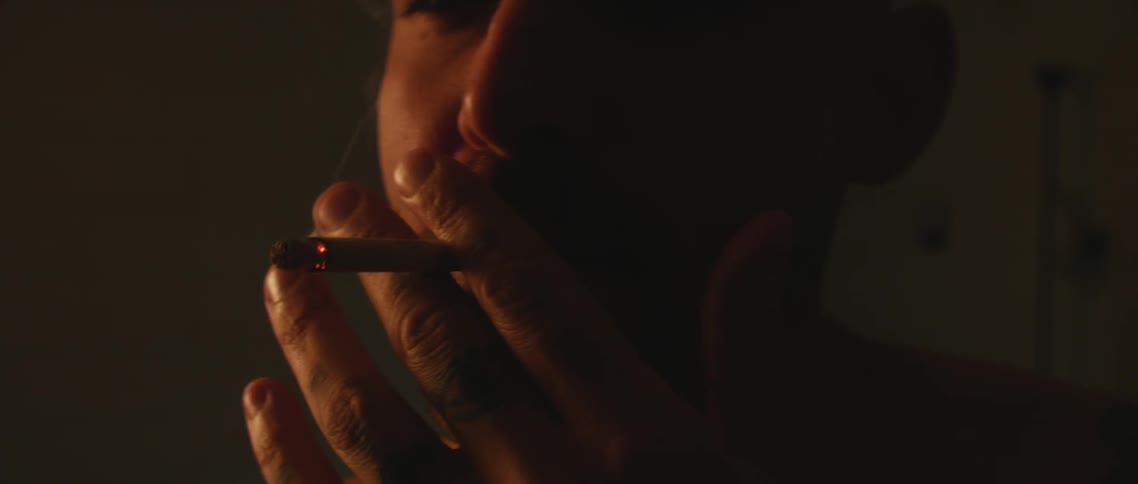 A mans fingers shake as he puffs a cigarette in a dimly lit room