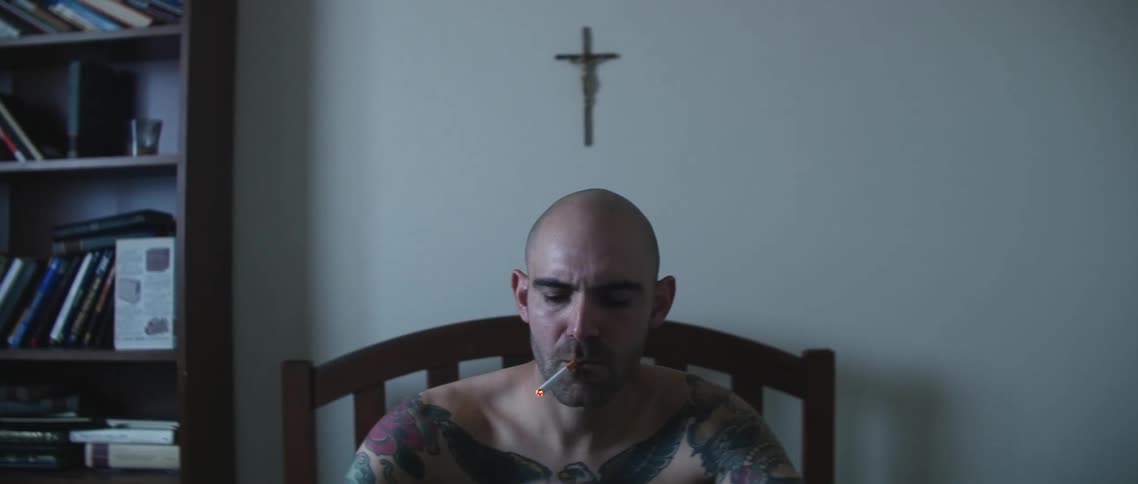 A man with tattoos over his chest and arms lights a cigarette and smokes
