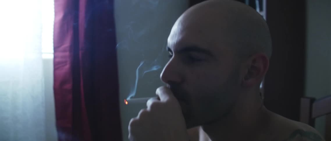 Tattooed man smoking a cigarette sitting in the bed on the bedroom at home