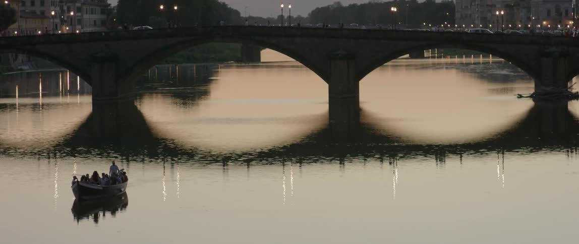 A tourist boat travelling along a river and traffic on a bridge in the background as night falls