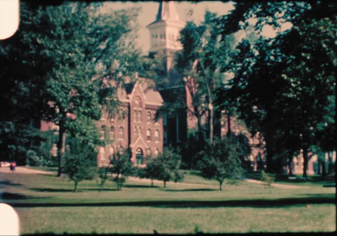 A car travels past a lush campus with a brick building and trees