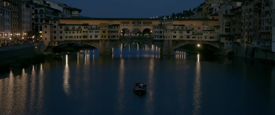 A boat filled with people makes its way along a river near an old city at night