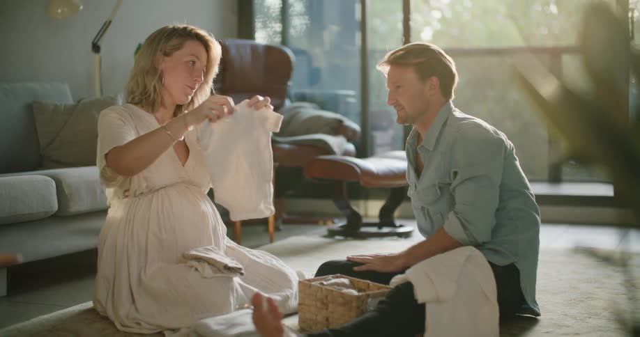 Pregnant couple sitting on the floor of the living room at sunset checking baby clothes