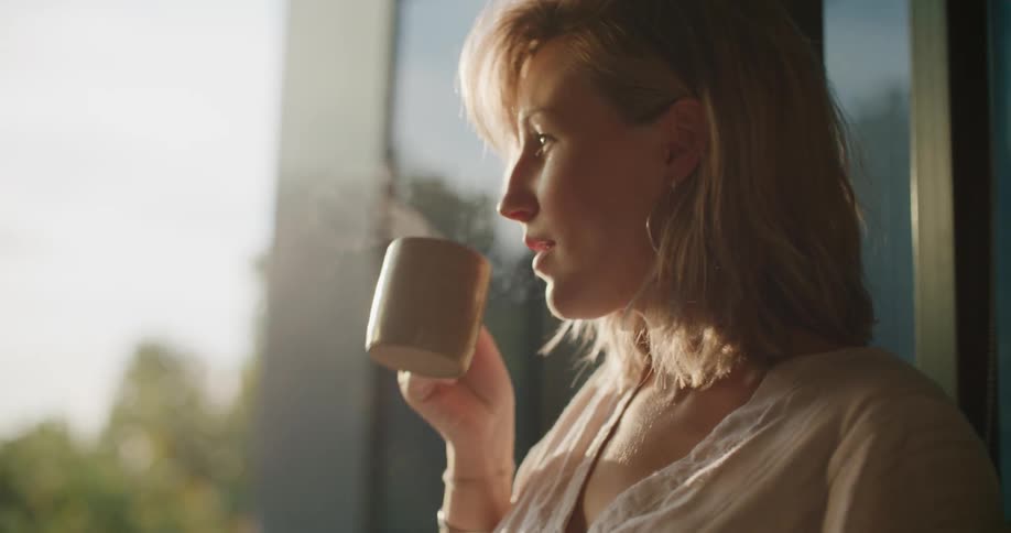 Pregnant woman holding her belly drinks a hot beverage near the window at sunset
