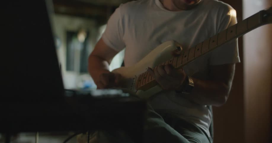A man wearing headphones plays a guitar while sitting indoors in front of a laptop