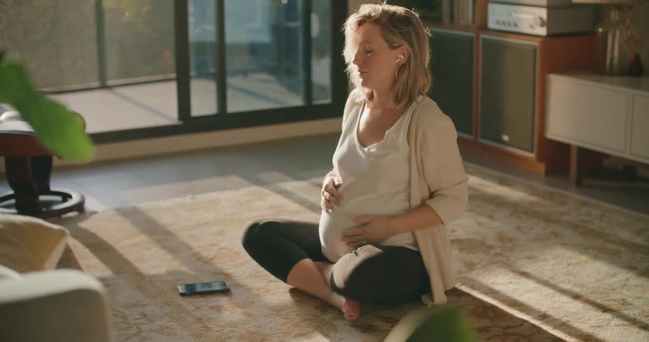 A pregnant woman sitting cross legged on the floor puts her ear buds in and takes a deep breath as she holds her belly