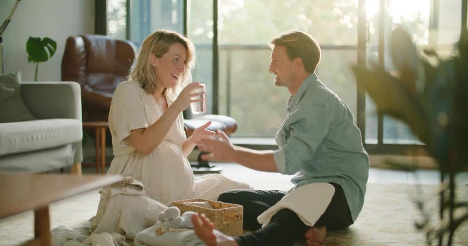 Pregnant woman and husband sit on rug in living room drinking beverages