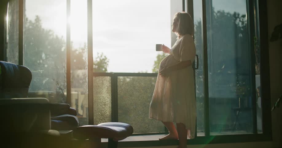 Blonde pregnant woman with white dress stands and drinks a cup of coffee