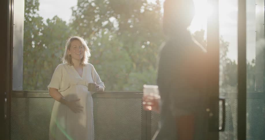 A pregnant woman standing on a balcony talking to a man with trees the sky and clouds in the background