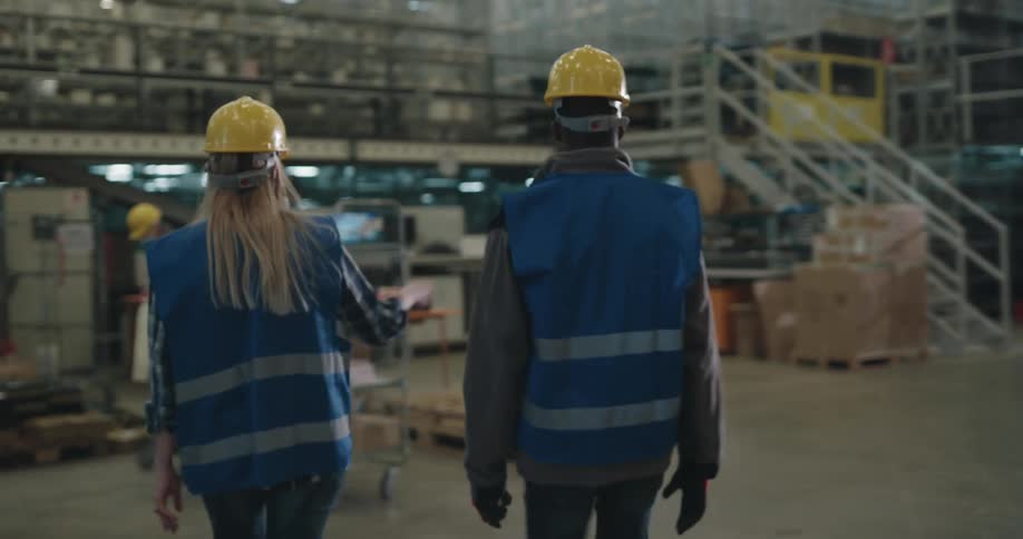 A man and a woman with hardhats and vests walking in a warehouse and waving with shelves in the background