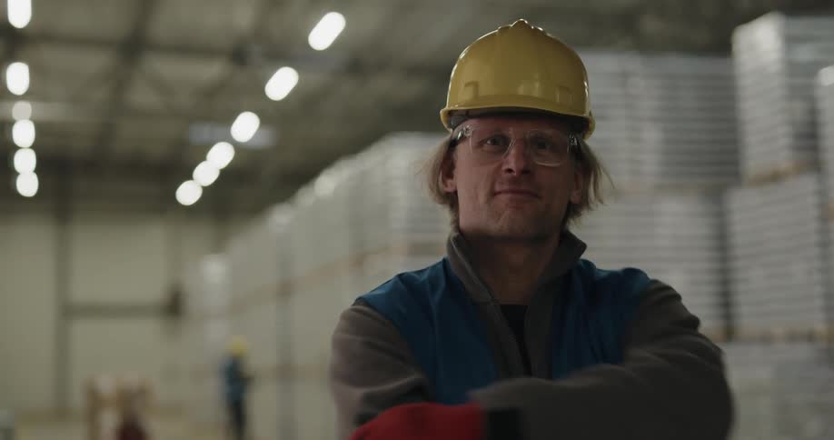 A man standing in a warehouse puts on a hard hat as he smiles into the camera