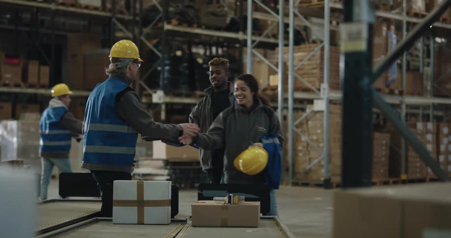 A man greets two smiling coworkers as they approach the conveyor and put on their hardhats