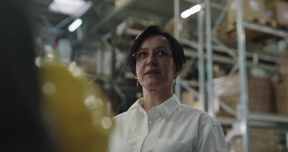 A woman in business attire stands in a warehouse talking to a worker holding his hardhat