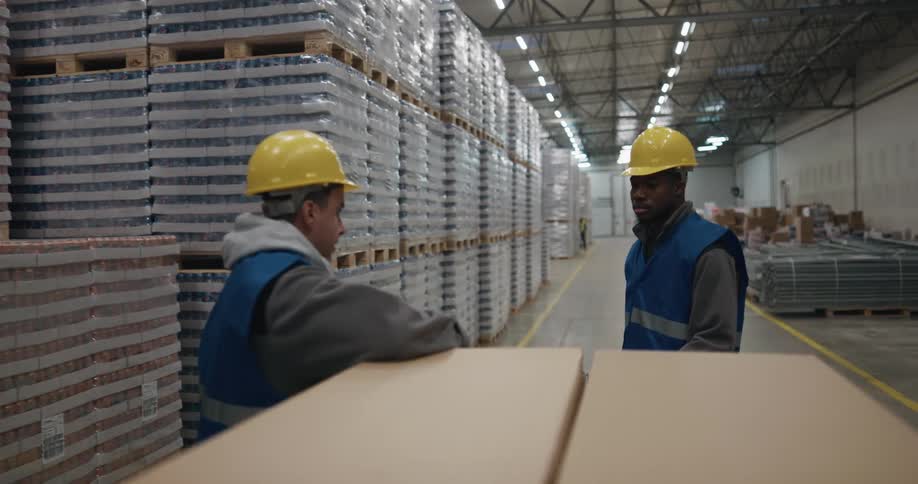 A man with a hardhat in a warehouse pulling boxes on a forklift and stopping to talk to another man