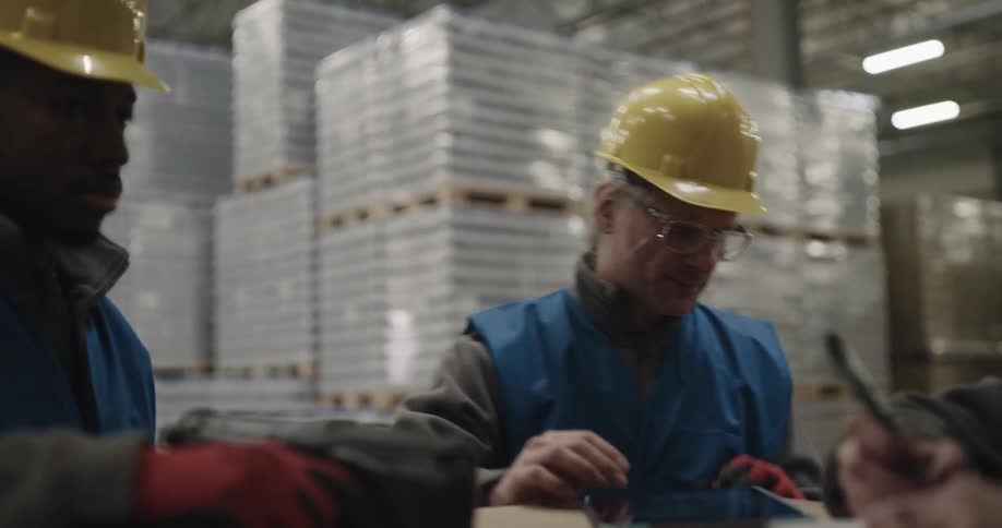 Three people in a warehouse full of boxes and pallets writing and speaking to each other