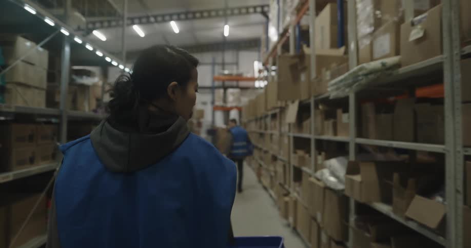 A woman carrying a basket walks through a warehouse looking at the boxes on shelves
