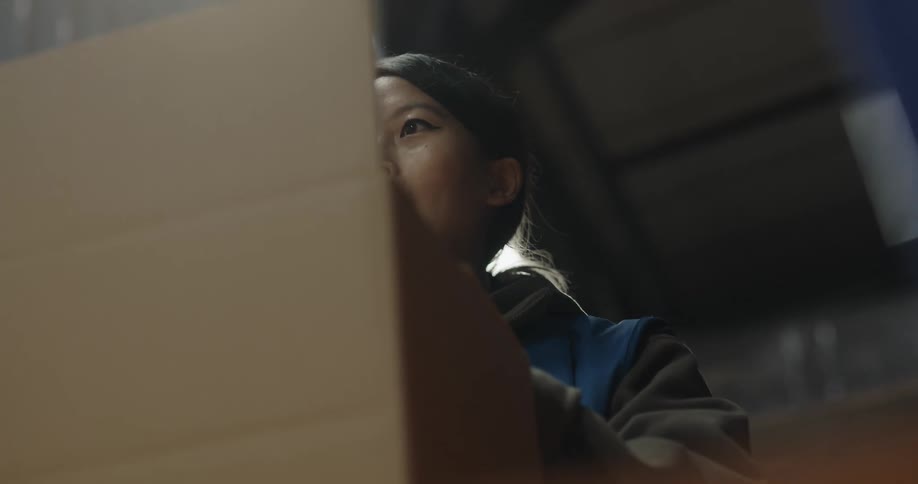 A woman with a vest in a warehouse folding a box with lights in the background