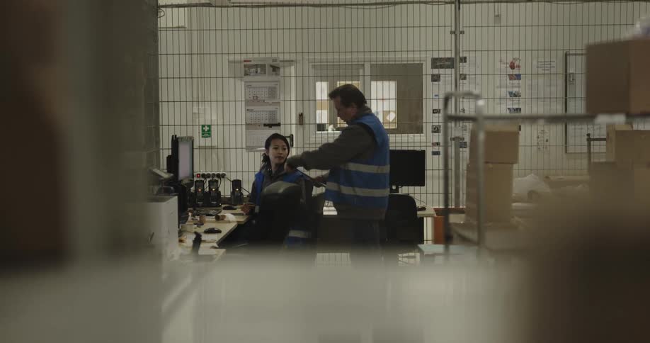 A man walking up to a woman sitting at a desk in front of a computer
