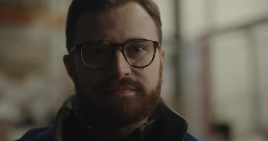 Bearded warehouse worker wearing glasses looks up at the camera and smiles